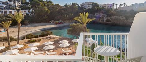 Un balcon avec une piscine, des chaises blanches et des parasols, donnant sur une plage et la mer.