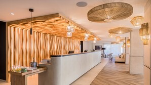 A modern reception area with a curved counter, wooden wall paneling, and multiple hanging light fixtures. - Barceló Ponent Beach (Felanitx)