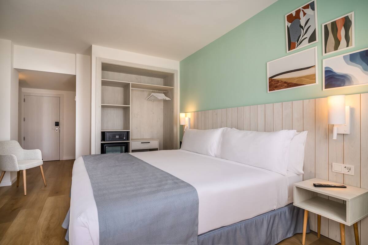 A hotel room with a large bed, a bedside table, a chair, a wardrobe, and artwork on the wall.
