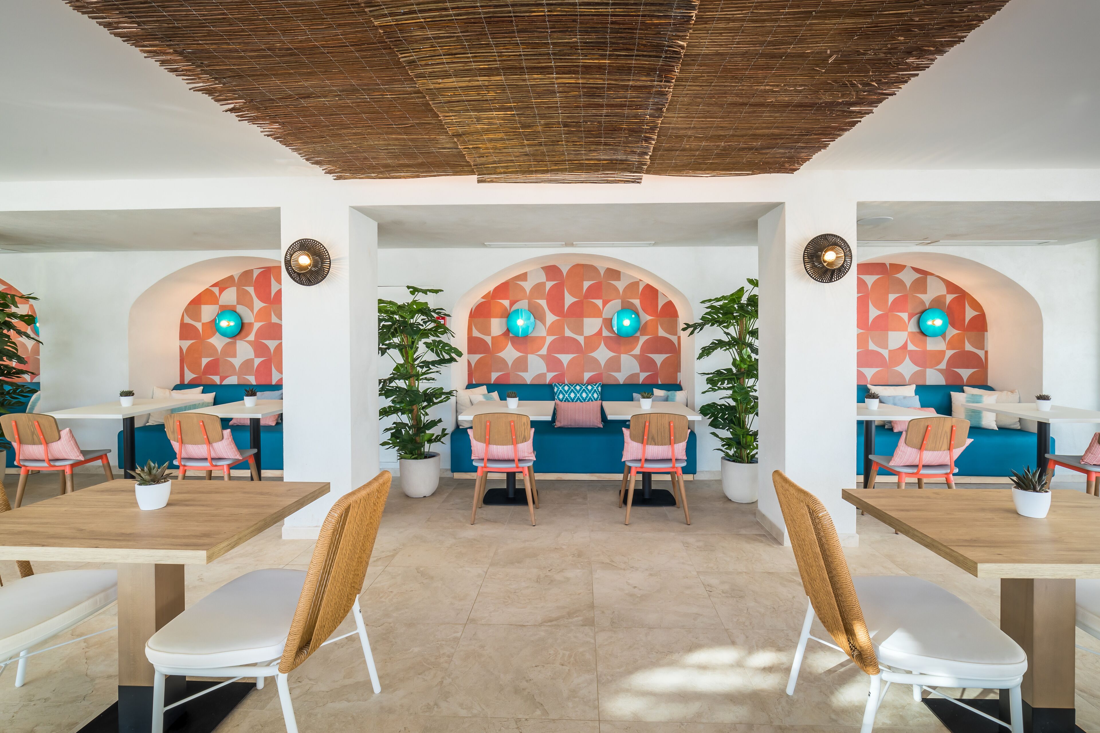 Un comedor moderno con mesas y sillas de madera, un muro con diseño y plantas en macetas.