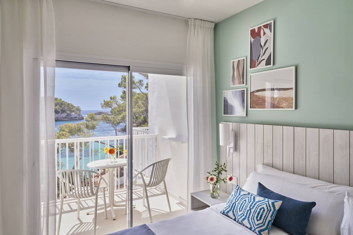 A hotel room with a balcony, a bed with blue and white pillows, a small table with chairs, and artwork on the wall.