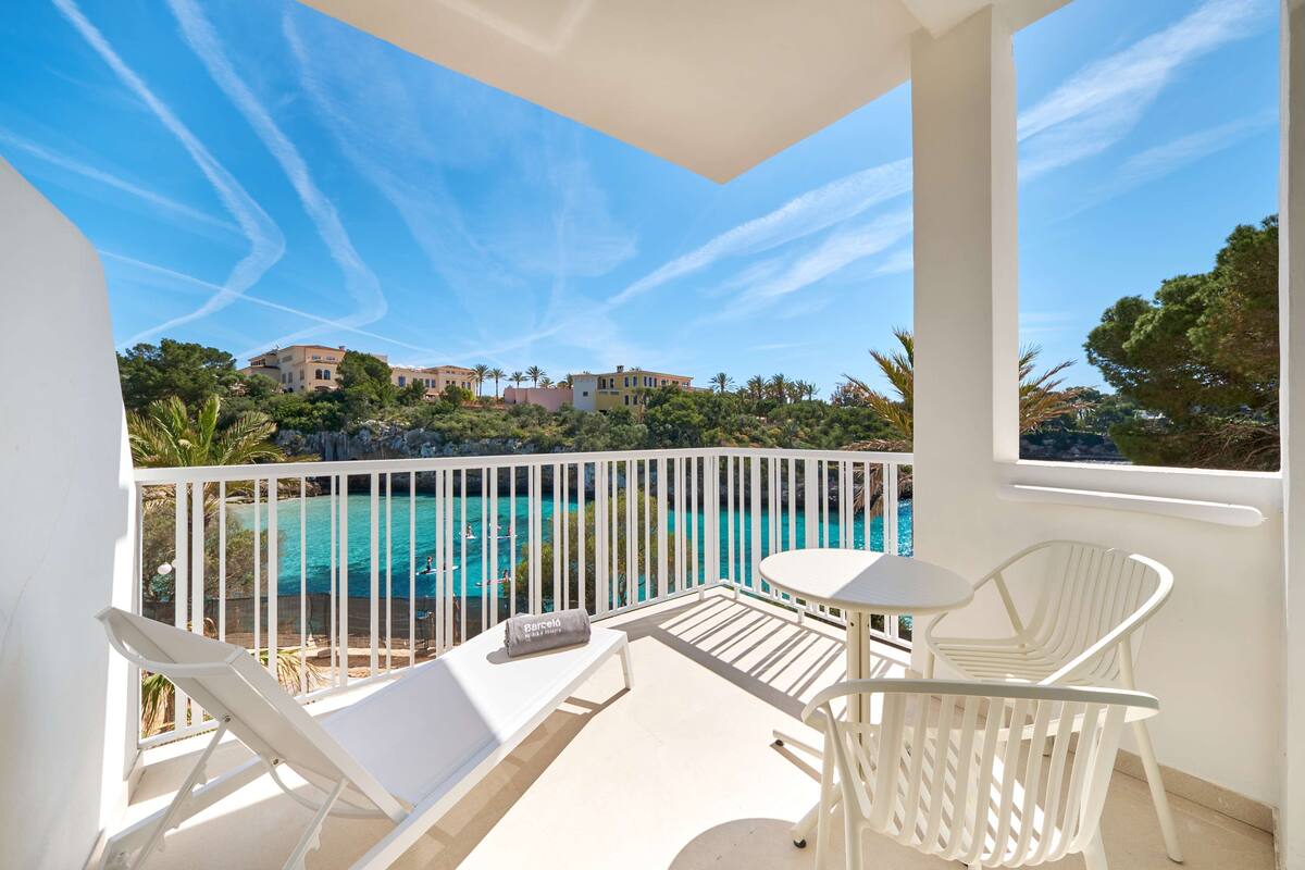 A balcony with white furniture, a view of a turquoise pool, and buildings in the background.