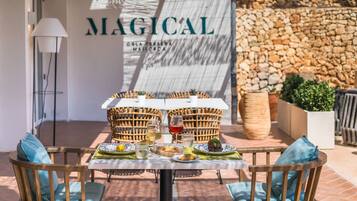 An outdoor dining area with wicker chairs and a table set with plates, glasses, and a bottle of wine. A stone wall and a lamp are visible in the background.