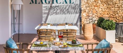 Un espace extérieur aménagé pour les repas, avec des chaises en osier et une table dressée avec des assiettes, des verres et une bouteille de vin. On aperçoit un mur de pierre et une lampe en arrière-plan.