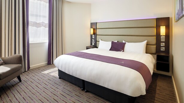 A hotel room with a large bed, a chair, a nightstand, and a wall-mounted lamp. - Premier Inn London Kensington (London)