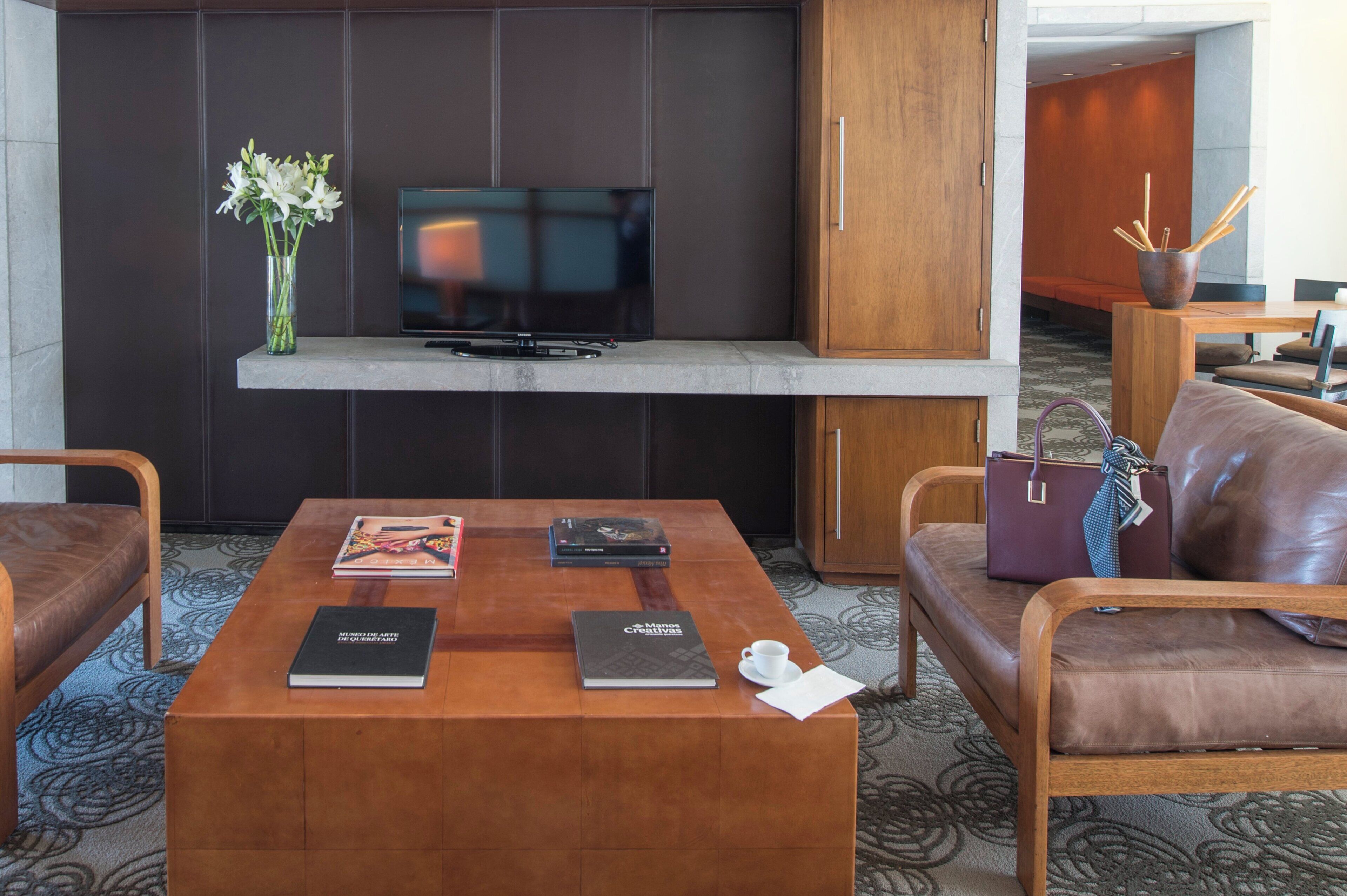 A modern hotel room with a flat-screen TV mounted on a dark wall, a wooden coffee table with books and a cup, leather sofas, and a wooden cabinet.