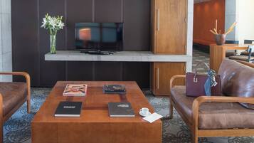 A modern hotel room with a flat-screen TV mounted on a dark wall, a wooden coffee table with books and a cup, leather sofas, and a wooden cabinet.