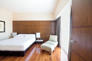 A hotel room with a large bed, a smaller bed, a nightstand with a lamp, a seating area, and a wardrobe.