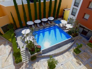 A rooftop pool with lounge chairs and umbrellas, surrounded by greenery and a tiled patio.