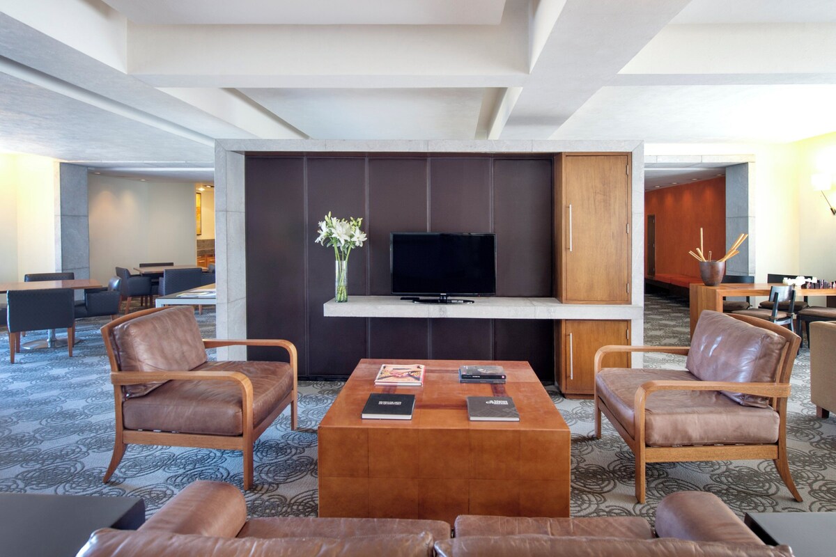 Un moderno vestíbulo de hotel con una zona de estar central que incluye sillones de cuero y una mesa de centro, un televisor de pantalla plana montado en un panel oscuro, y un florero con flores blancas.