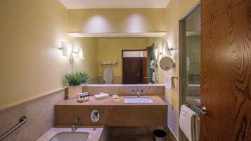 Un baño con bañera, lavabo y espejo. Hay un perchero, un cubo de basura y una planta en la encimera.