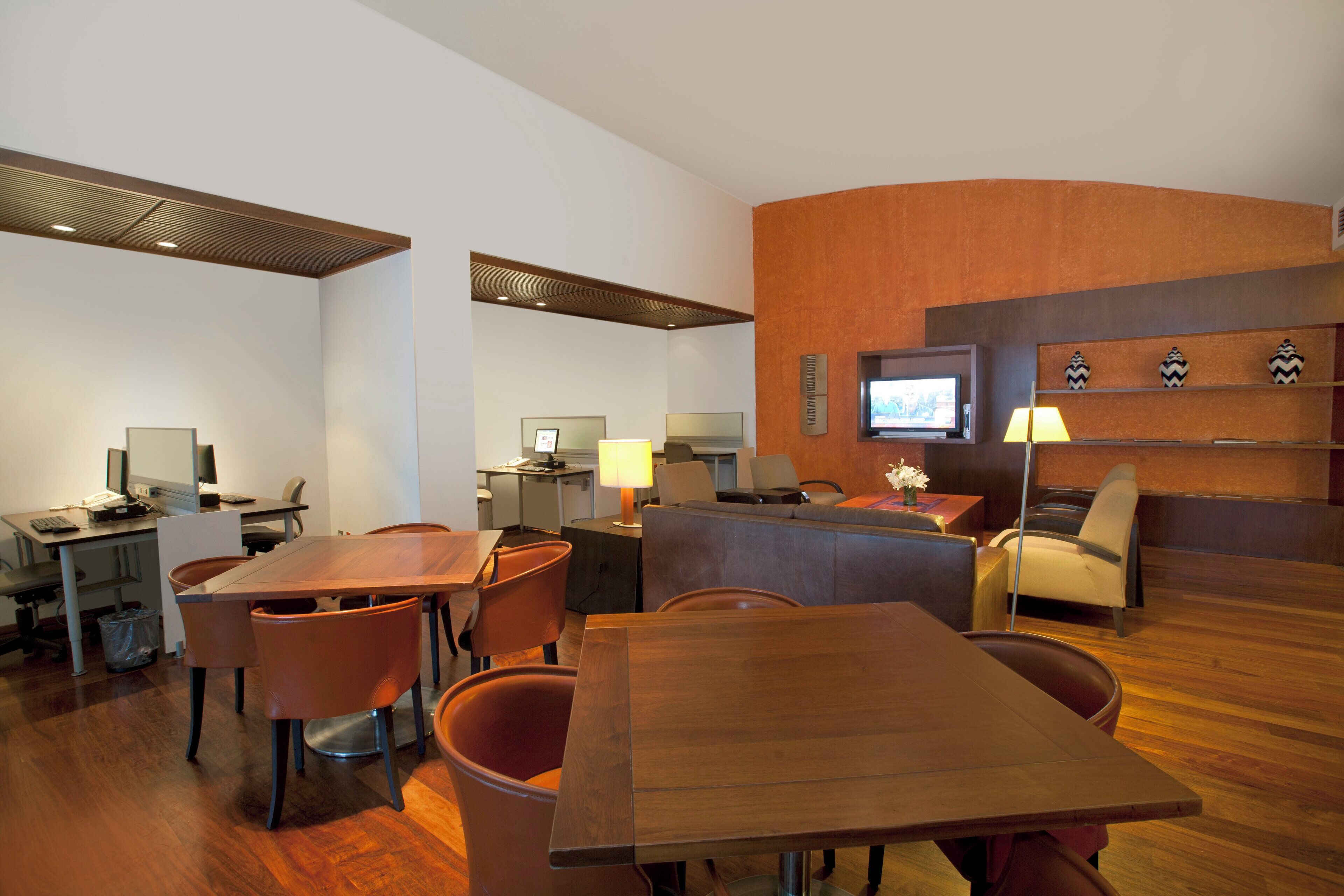 Una habitación de hotel moderna con una mesa de comedor de madera, asientos cómodos, un televisor y un escritorio con una computadora.