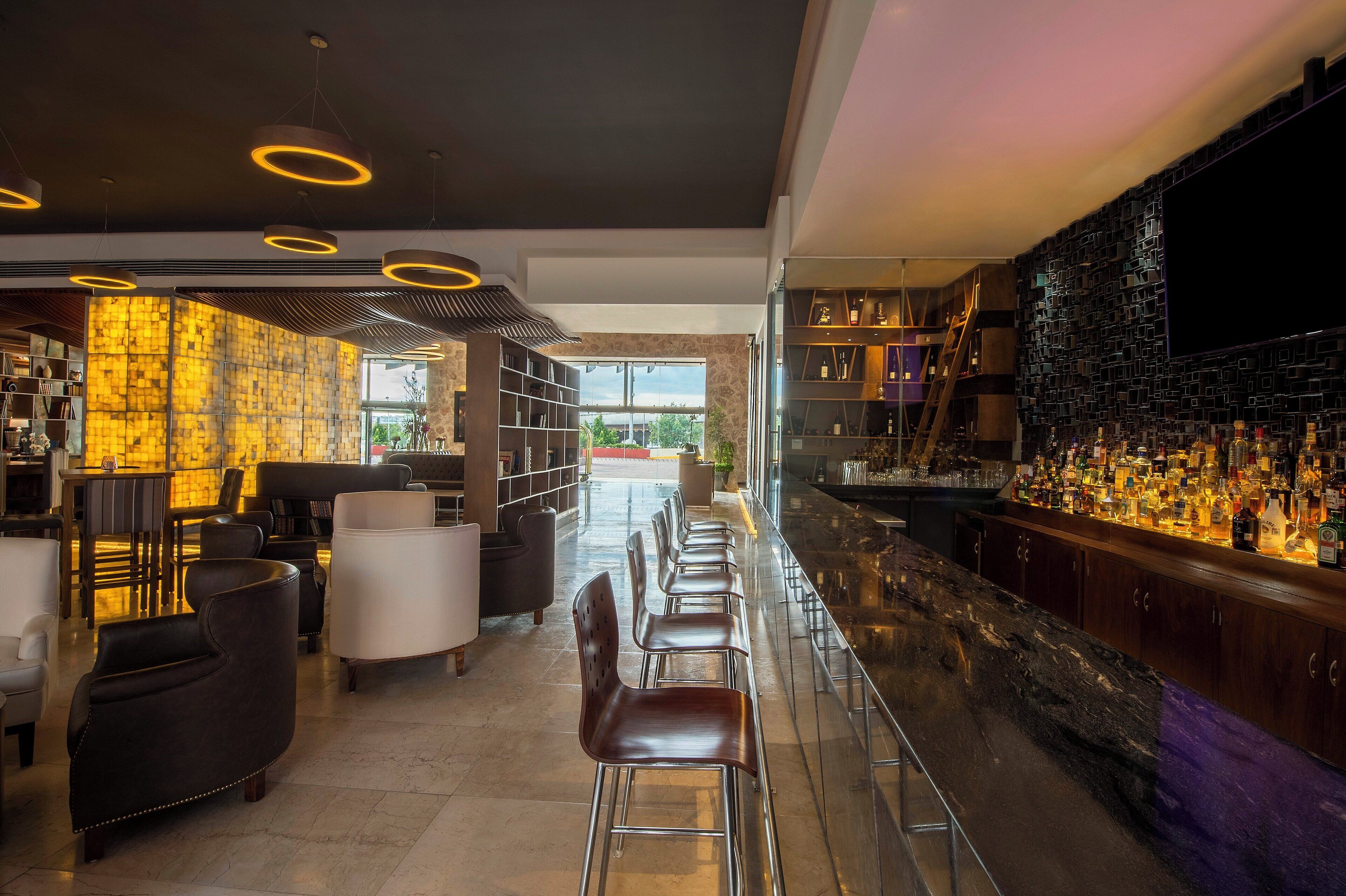 Un moderno espacio de bar con una barra larga, taburetes y una variedad de botellas. Hay estanterías y una zona de estar con sillas y un sofá.