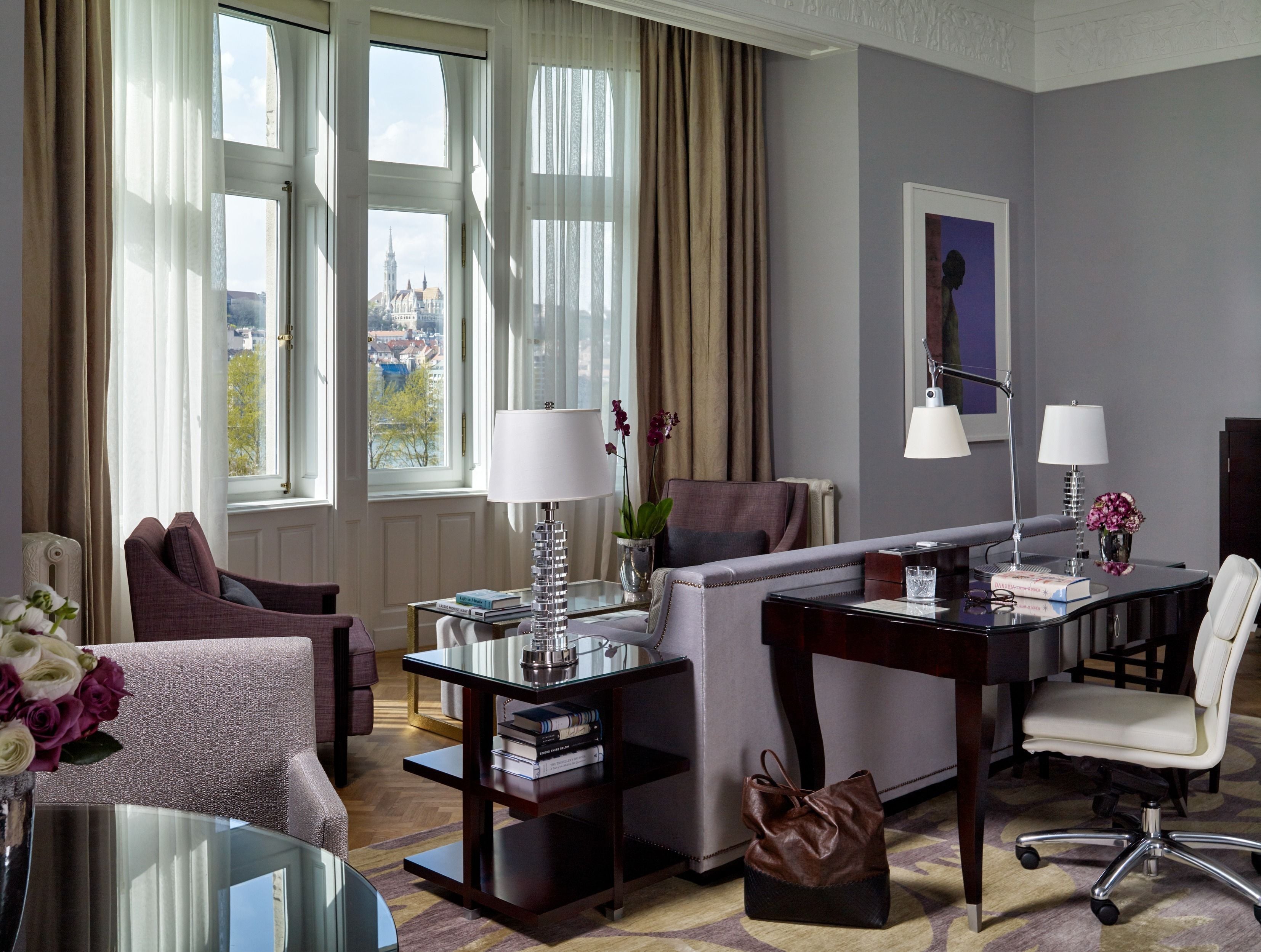 Quarto de hotel com sofá, poltrona, escrivaninha e cadeira. Possui duas janelas com cortinas, uma luminária, um vaso com flores e um quadro na parede.