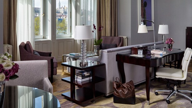 A hotel room with a sofa, armchair, desk, and chair. There are two windows with curtains, a lamp, a vase with flowers, and a framed picture on the wall. - Four Seasons Gresham Palace (Budapest)