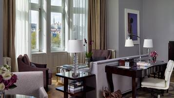Quarto de hotel com sofá, poltrona, escrivaninha e cadeira. Possui duas janelas com cortinas, uma luminária, um vaso com flores e um quadro na parede.