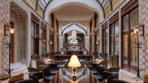 A spacious lobby with a high arched glass ceiling, a central round table with chairs, and a grand staircase in the background. - Four Seasons Gresham Palace (Budapest)
