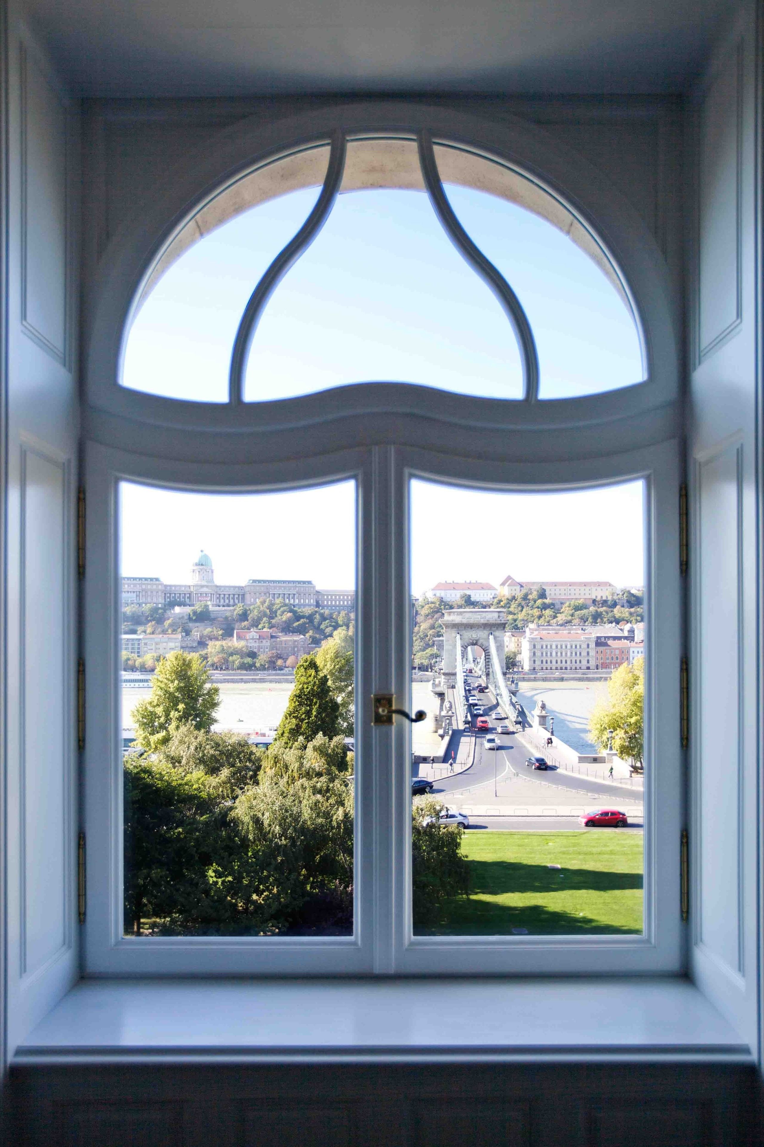 A view through a white-framed window with a curved top, overlooking a bridge, a river, and a cityscape with buildings and greenery.