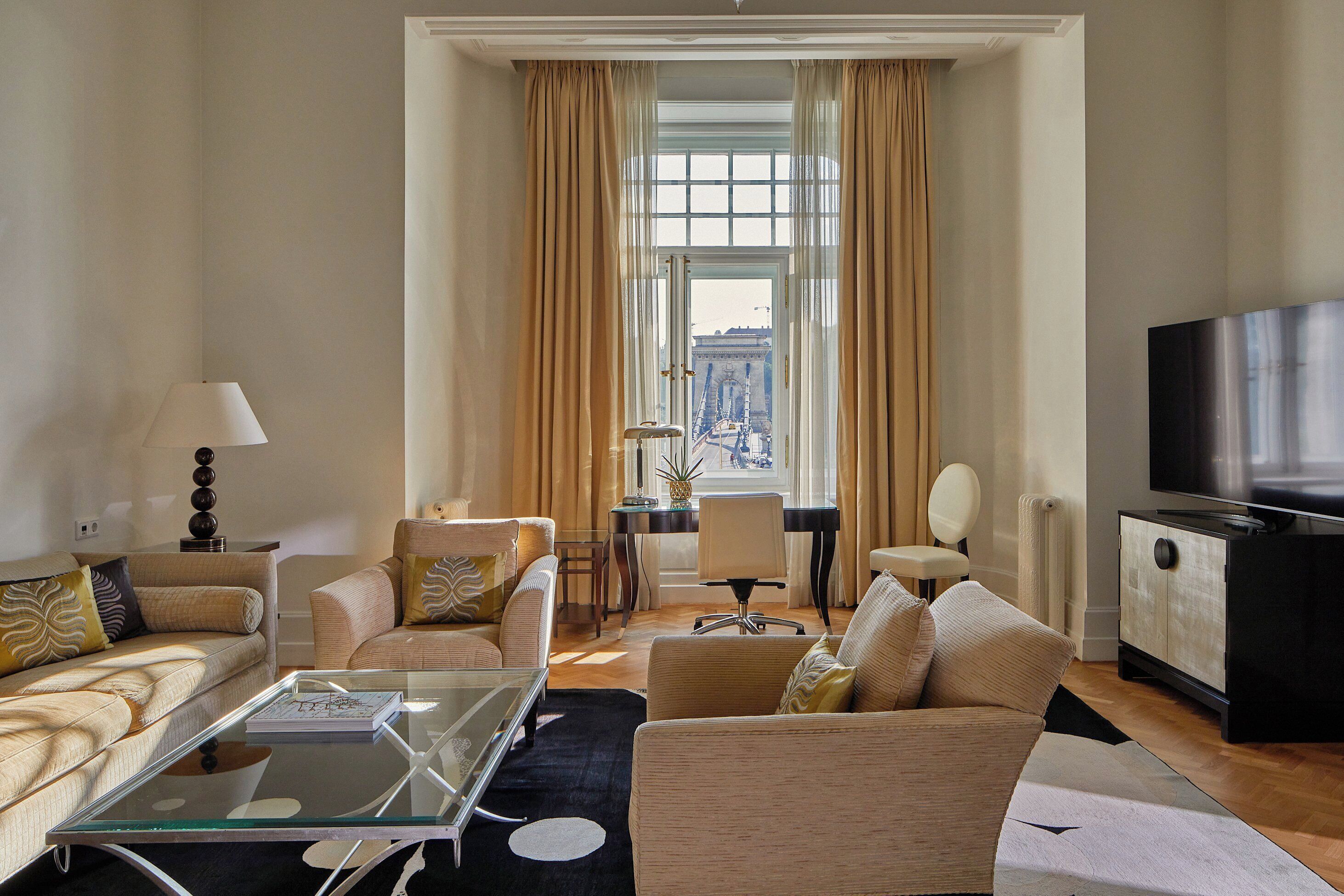 Una sala de estar moderna con un sofá, sillones, una mesa de centro de vidrio, un televisor y vistas a un puente por la ventana.