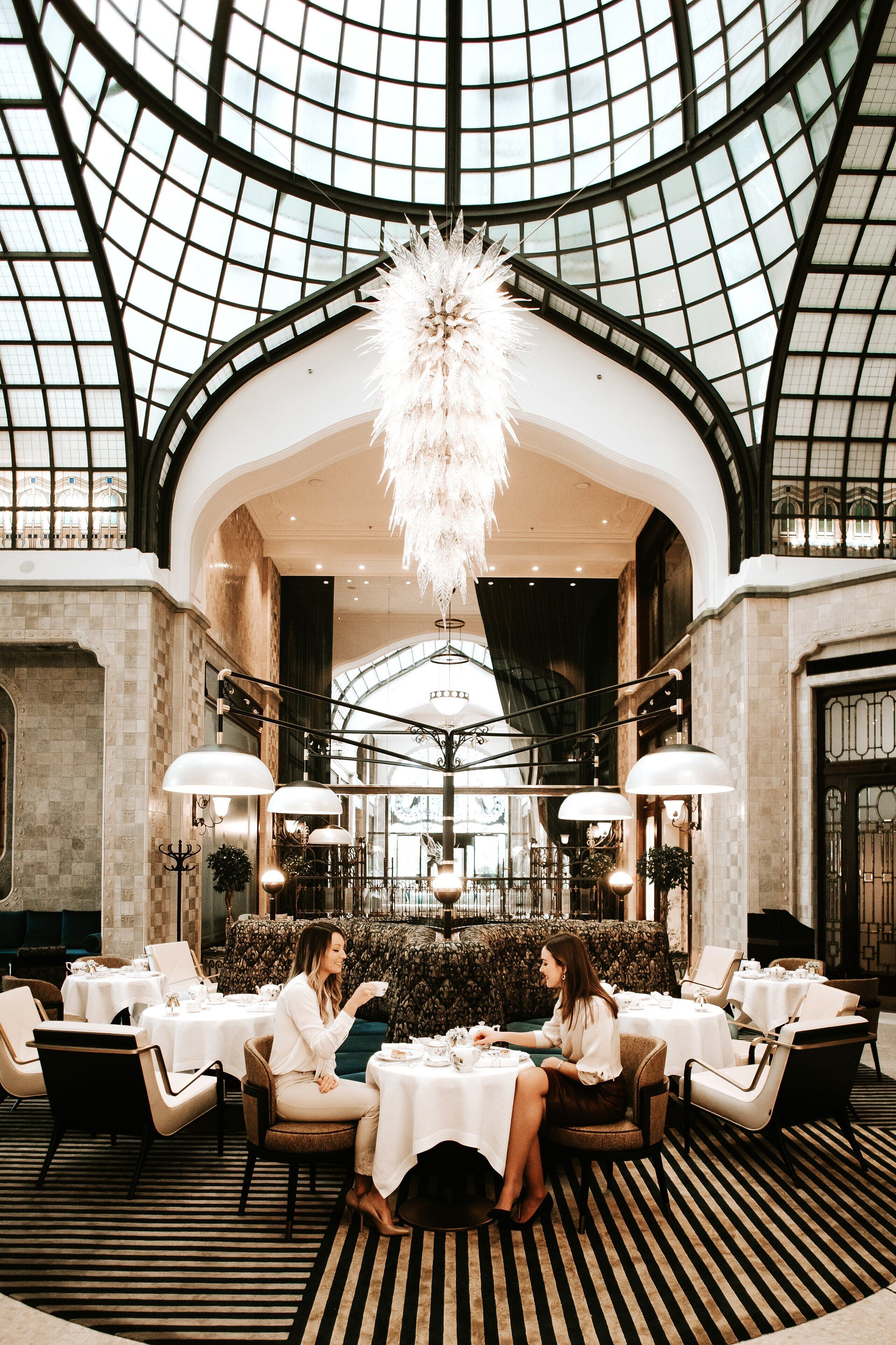 Un lobby de hotel lujoso con un techo alto, candelabros ornamentales y una zona de estar central con mesas y sillas.