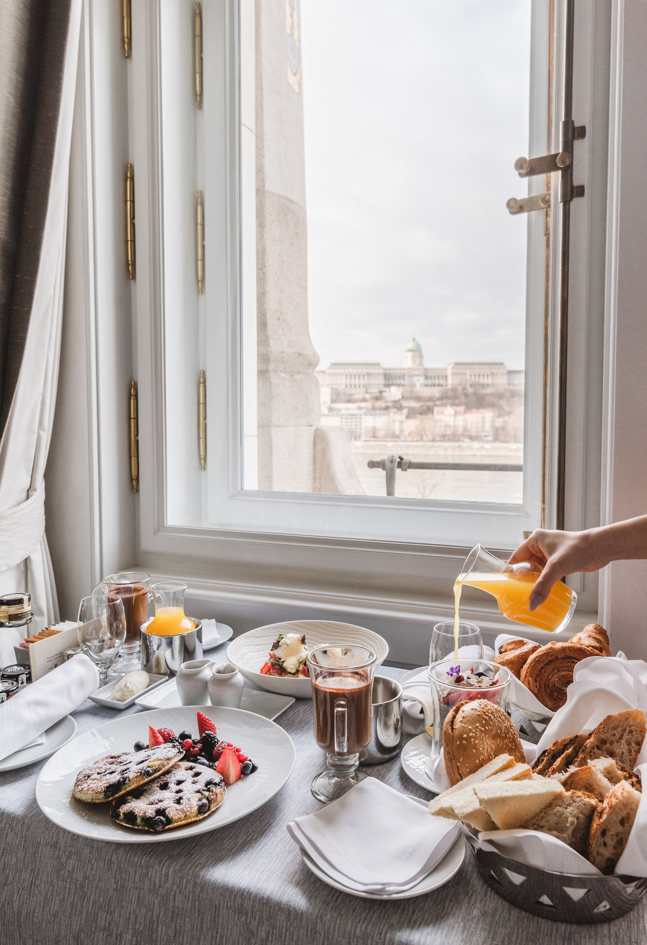 A breakfast table with pastries, fruits, and beverages, overlooking a cityscape.