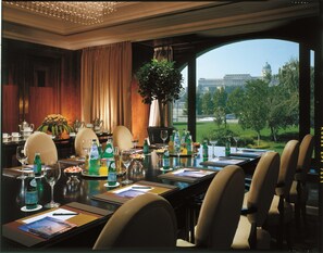 A formal dining room with a long table set for a meal, a large window offering a view of a building and greenery, and a chandelier hanging from the ceiling.