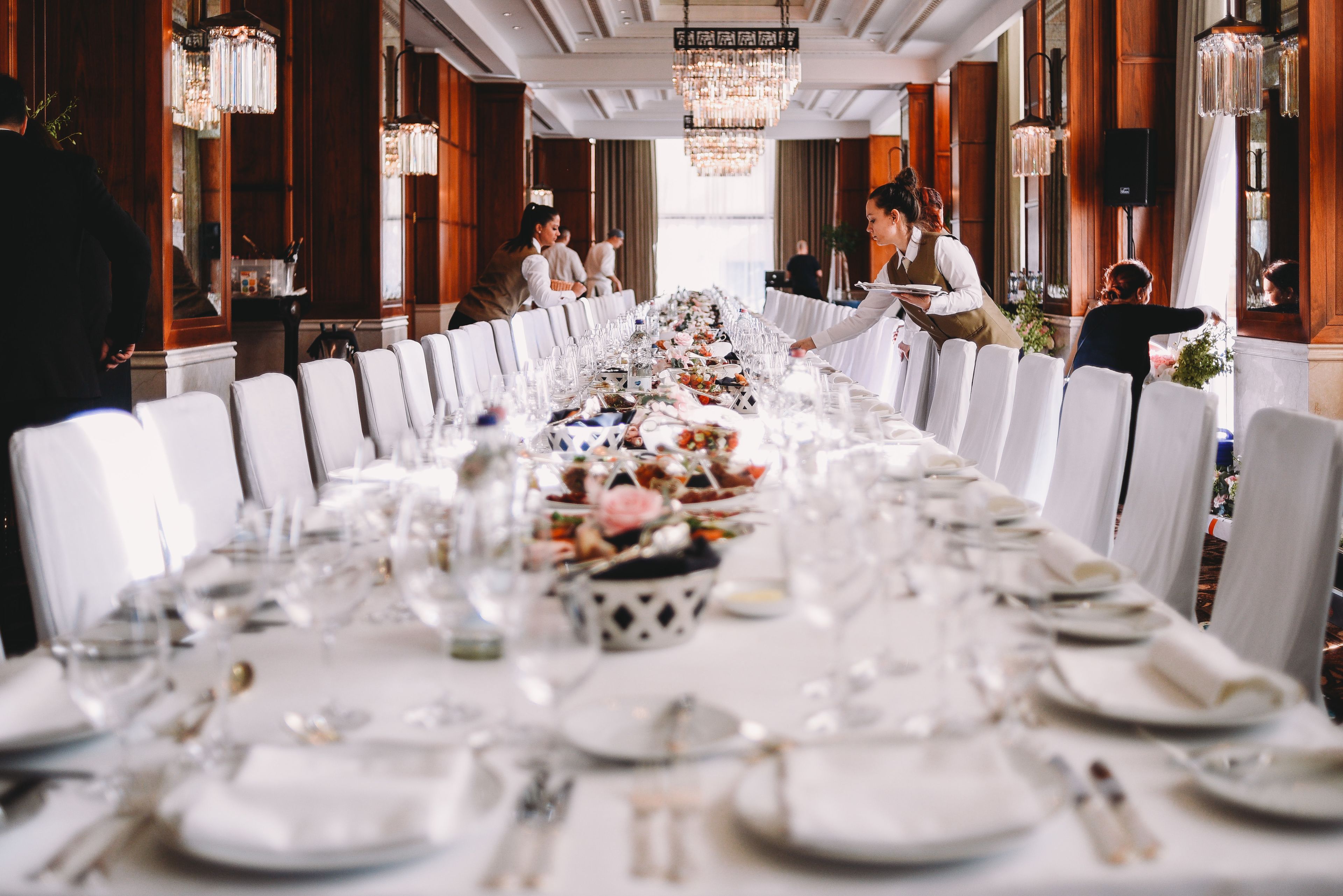 A long banquet table set with white tablecloths, plates, and glasses, surrounded by chairs.