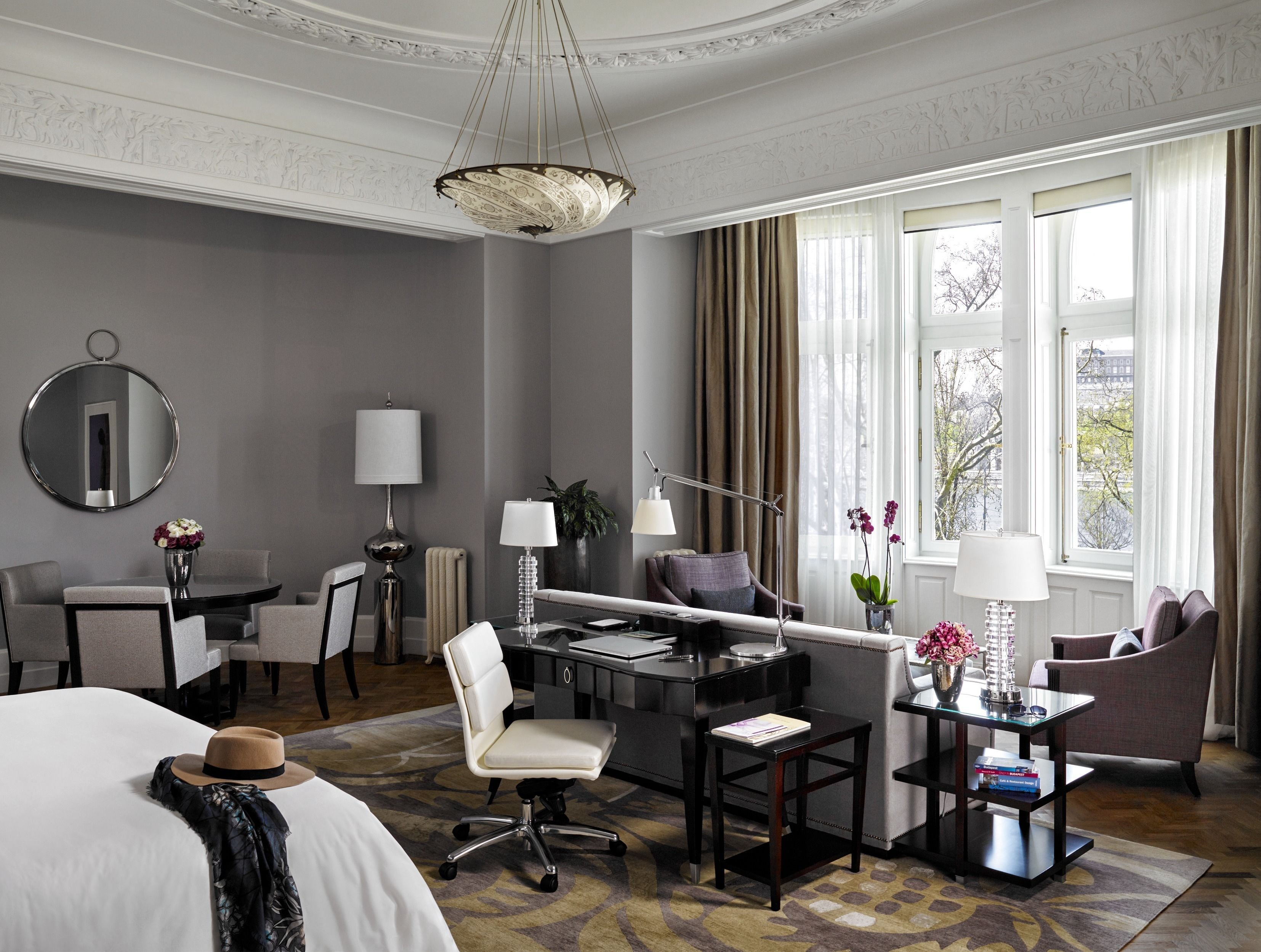 A hotel room with a bed, desk, chair, and a view of the city through large windows.