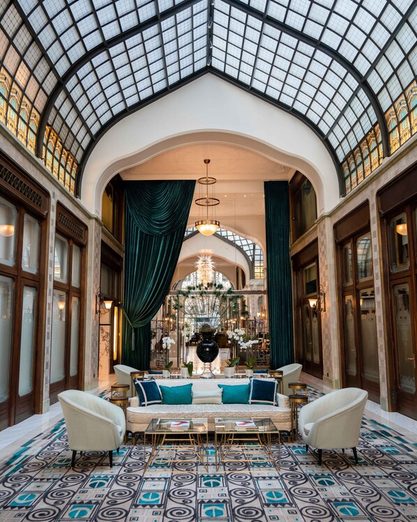 A grand lobby with a high arched glass ceiling, ornate wooden doors, and a central seating area with a patterned floor.