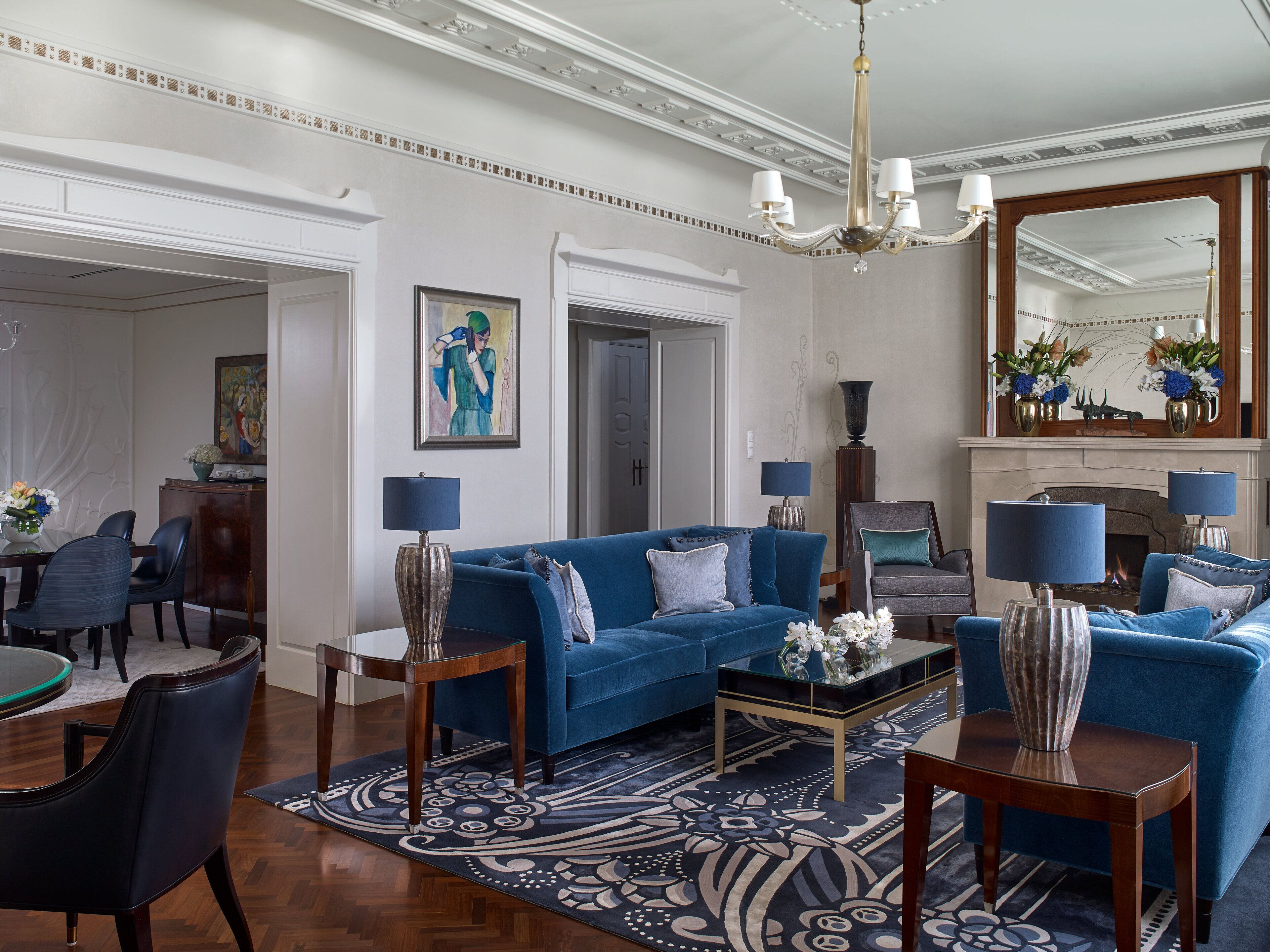 A spacious living room with a blue sofa, a coffee table, and a chandelier. There is a fireplace with a mirror above it and a dining area in the background.
