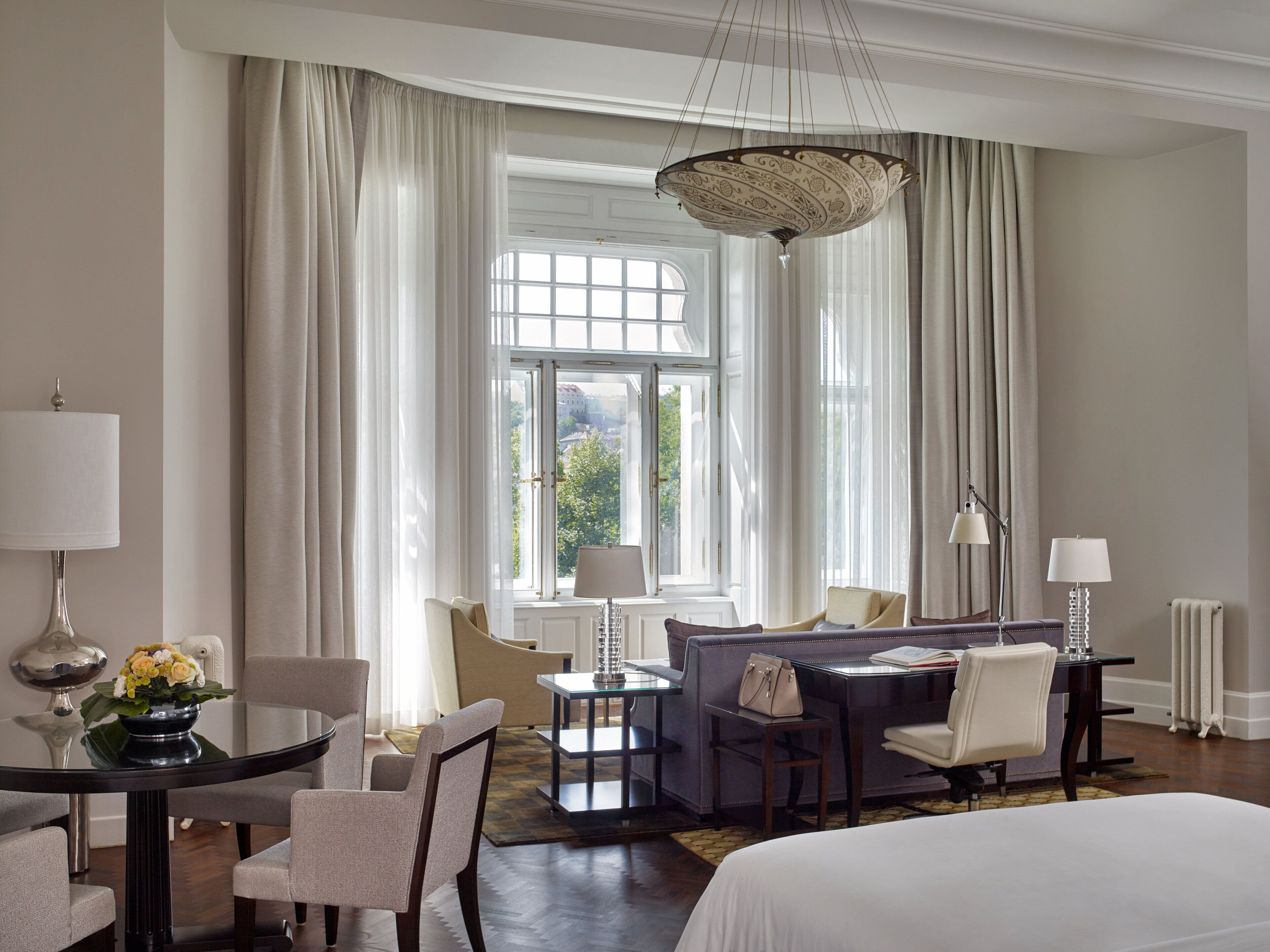 Habitación de hotel con una cama grande, una zona de estar con sofá y sillones, una mesa de comedor con sillas y vistas al exterior a través de amplios ventanales.