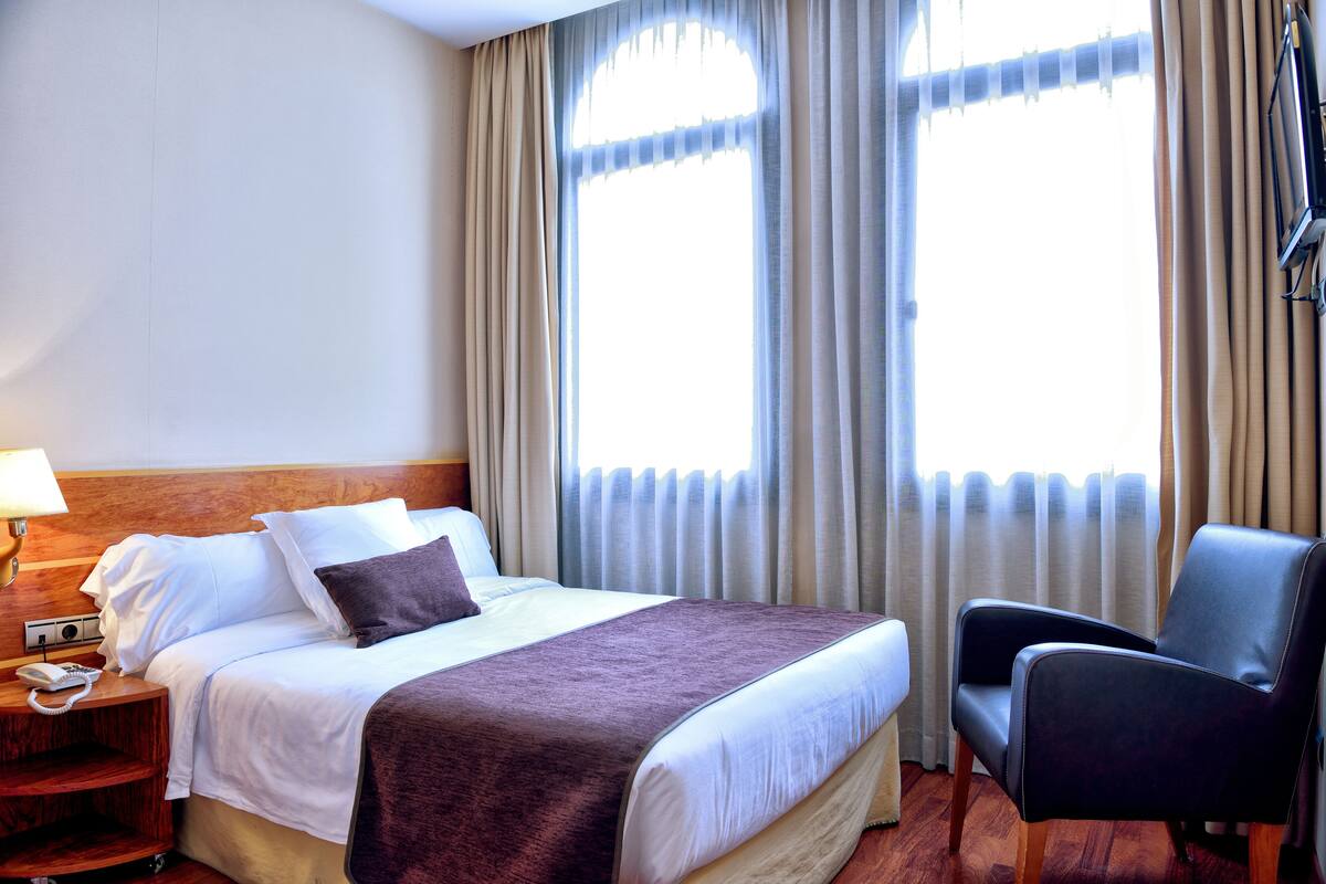 Een hotelkamer met een bed, een stoel, een nachtkastje met een telefoon en een raam met gordijnen.