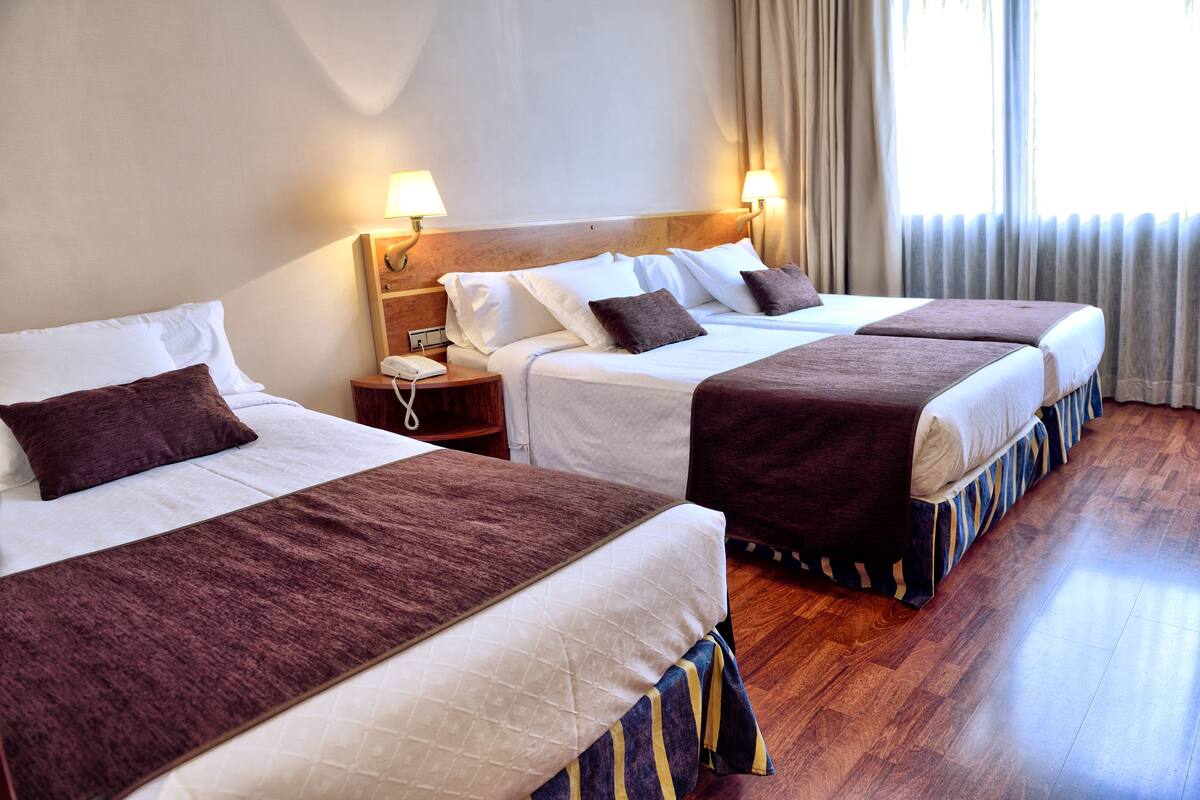 Hotelkamer met twee bedden, elk met een bruine plaid, witte lakens en een houten hoofdbord. Er staan nachtlampjes, een telefoon op het nachtkastje en een raam met gordijnen.
