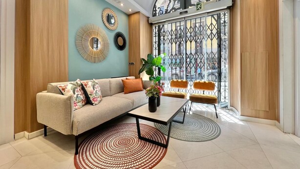 Uma sala de estar moderna com um sofá, mesa de centro e duas cadeiras. Há uma janela grande com portão de metal decorativo e vista para a rua.