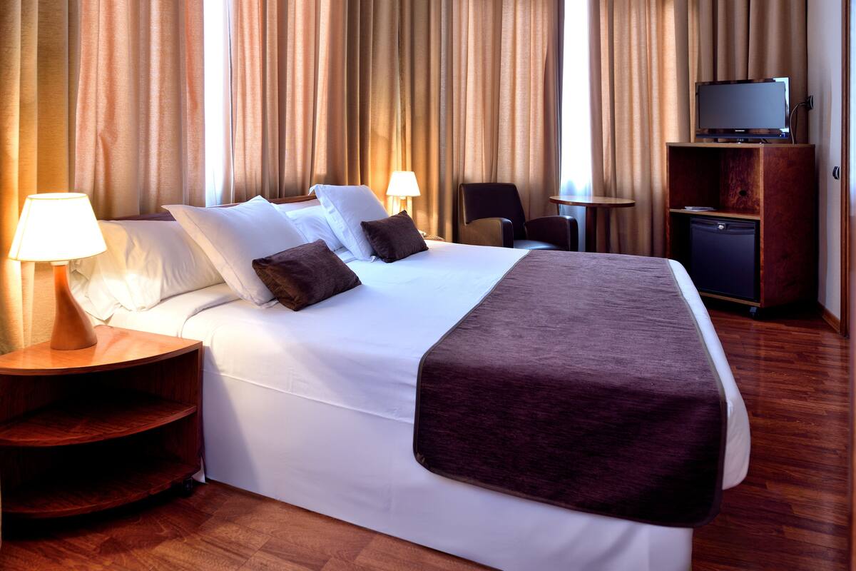 Een hotelkamer met een groot bed, twee nachtkastjes, een televisie op een houten kast en een klein bureau met stoel.