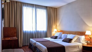 Quarto de hotel com duas camas, televisão, escrivaninha e janela com cortinas.