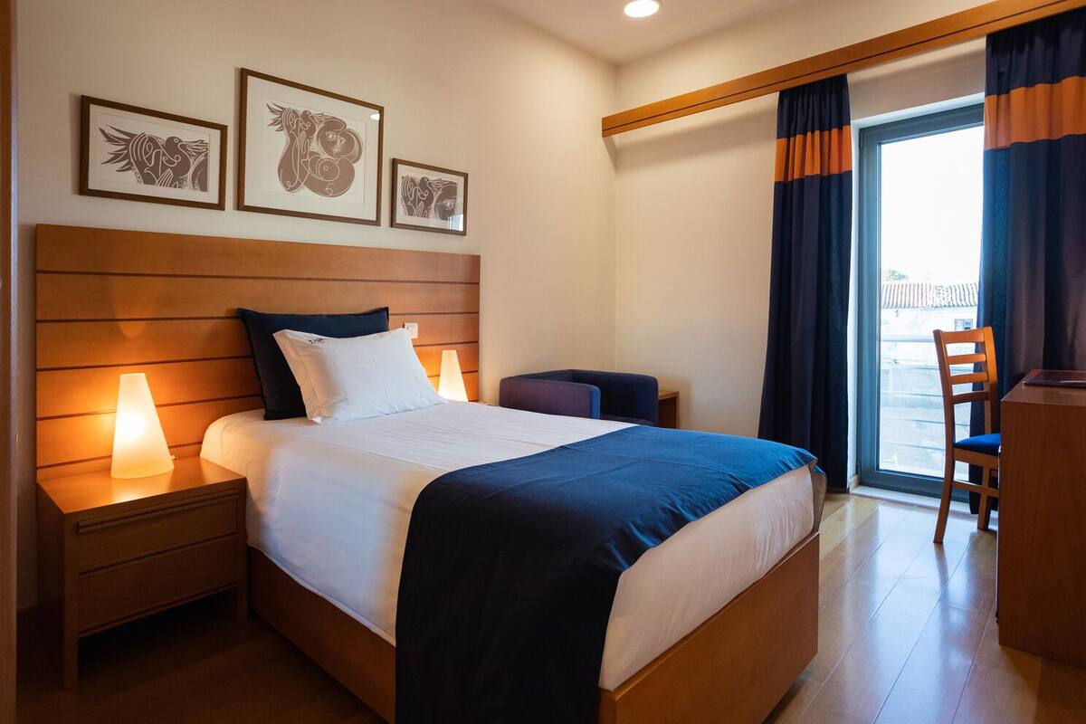 A hotel room with a large bed, bedside lamps, a desk, and a chair. There are three framed artworks on the wall above the bed.