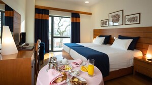 A hotel room with a large bed, a bedside table with a lamp, a desk with a lamp, a window with curtains, and a breakfast table with food and drinks. - Hotel Pombalense (Pombal)