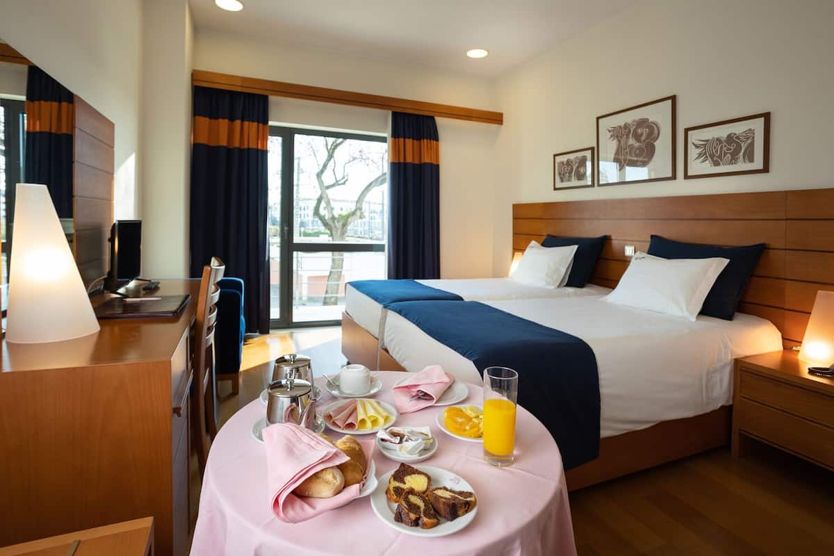 A hotel room with a large bed, a bedside table with a lamp, a desk with a lamp, a window with curtains, and a breakfast table with food and drinks.