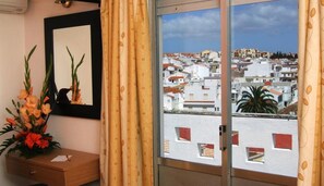 A room with a view of a cityscape through a glass door, a mirror on the wall, and a wooden side table with a flower arrangement. - Riomar (Lagos)