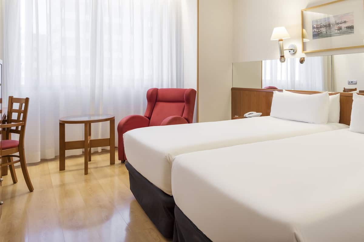 A hotel room with two beds, a red armchair, a wooden desk, and a television.