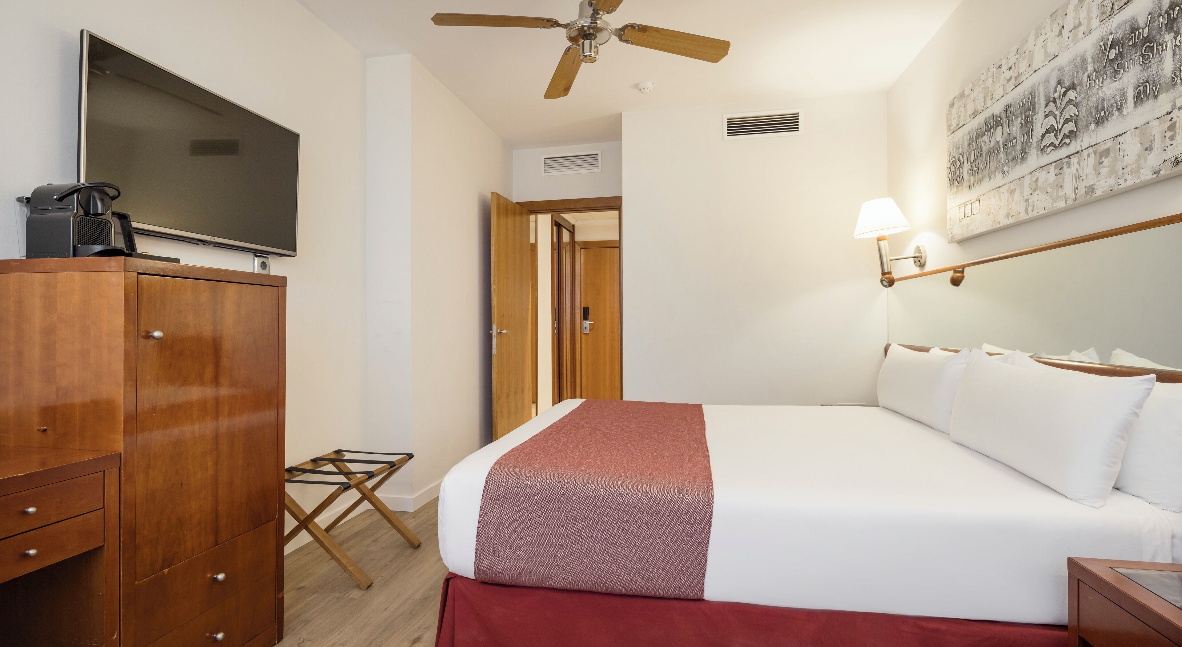 Een hotelkamer met een bed, een televisie op een houten kast, een plafondventilator en een wandlamp.