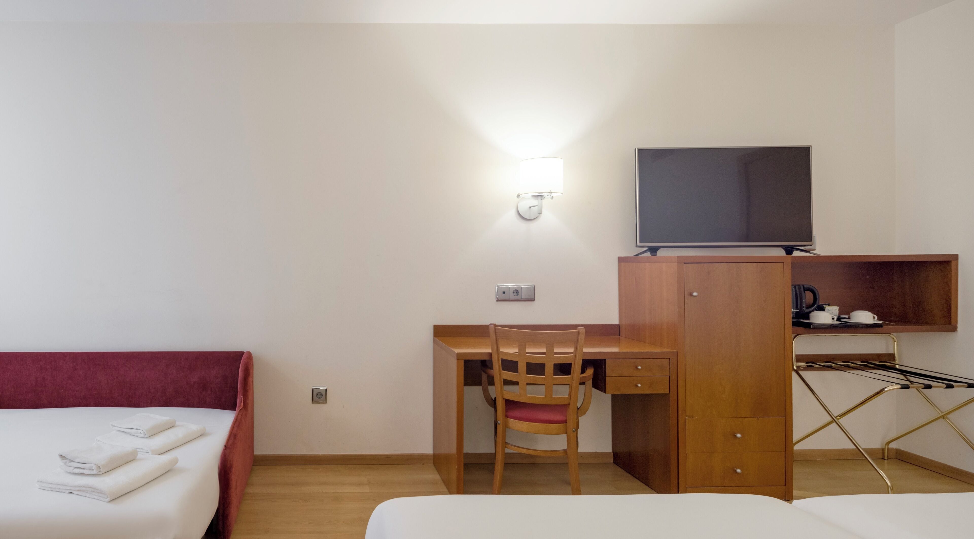 Quarto de hotel com duas camas, uma escrivaninha com cadeira, televisão, prateleira com chávenas e chaleira, e luminária na parede.