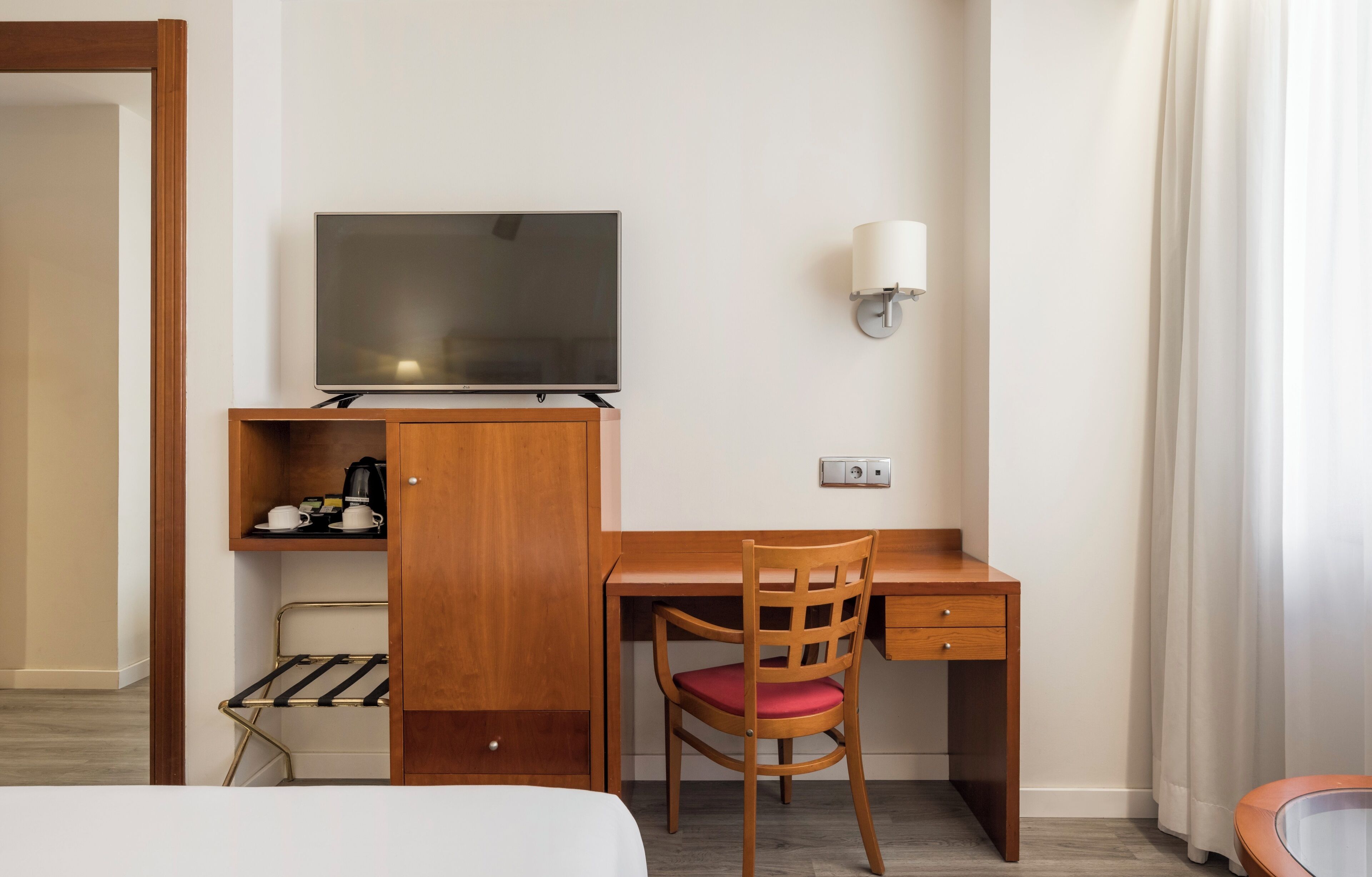 A hotel room with a wooden desk, a television, a chair, a lamp, and a bed.