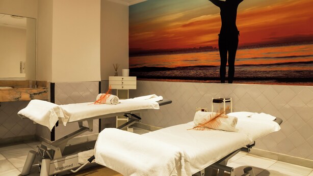 A medical examination room with two treatment beds, a marble-tiled counter, and a large painting of a person with arms raised against a sunset backdrop.