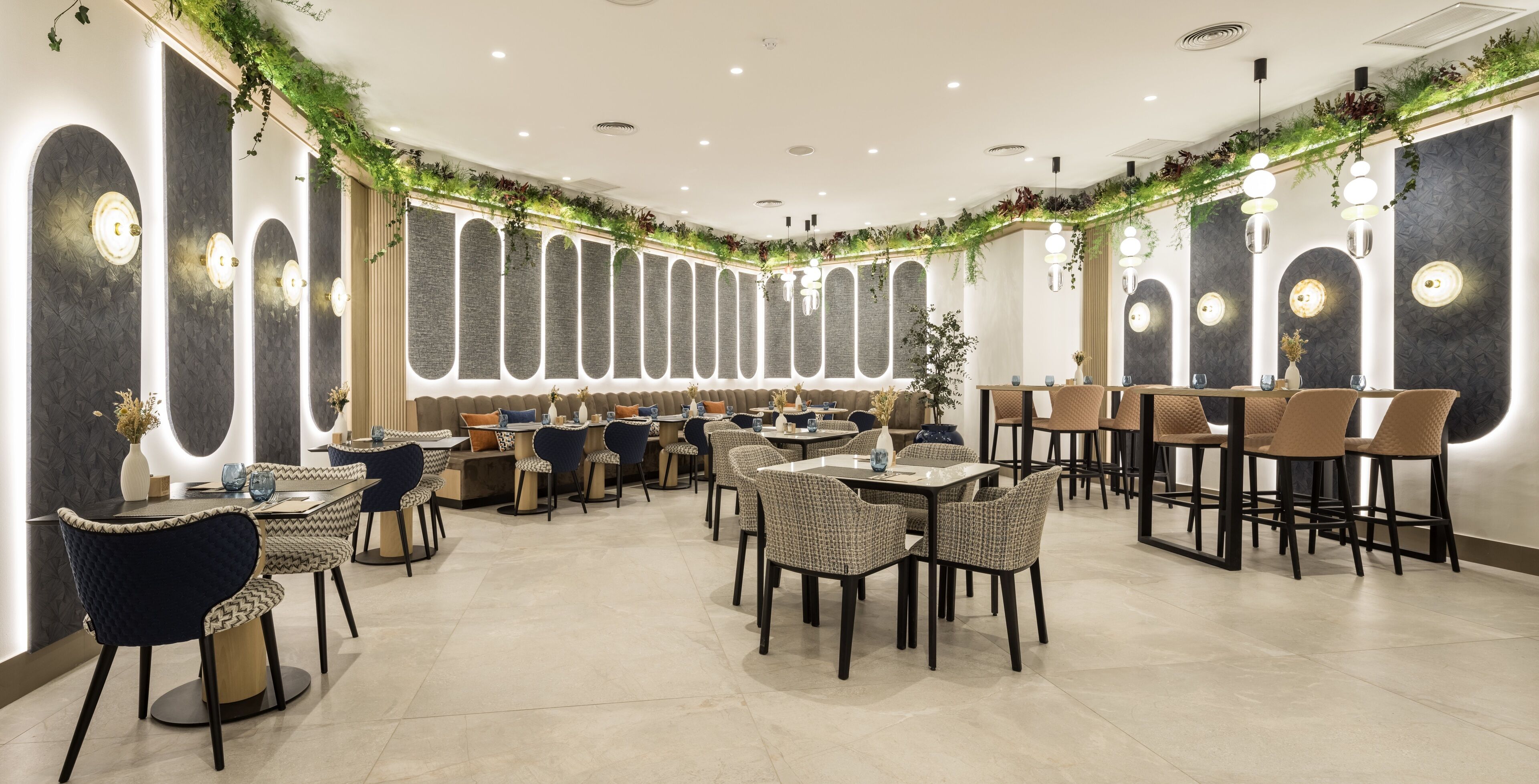 A modern restaurant with round tables and chairs, decorative wall lighting, and greenery accents.