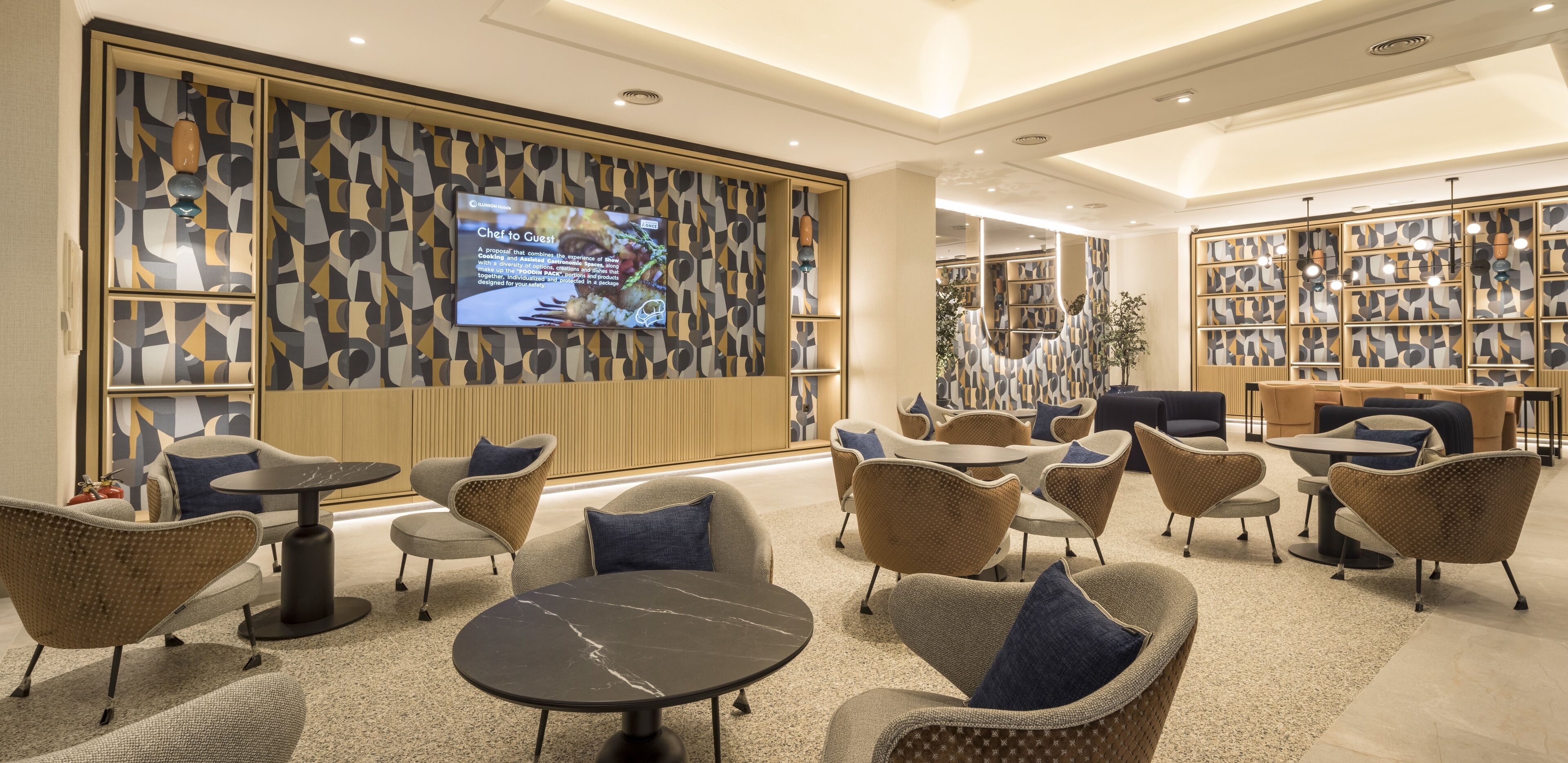 A modern lounge area with comfortable seating, a large screen displaying content, and a patterned wall.