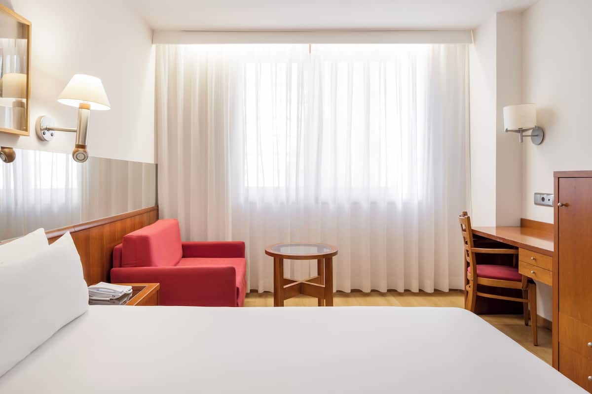 A hotel room with a bed, a red armchair, a small table, a desk with a chair, a TV, and a wardrobe.