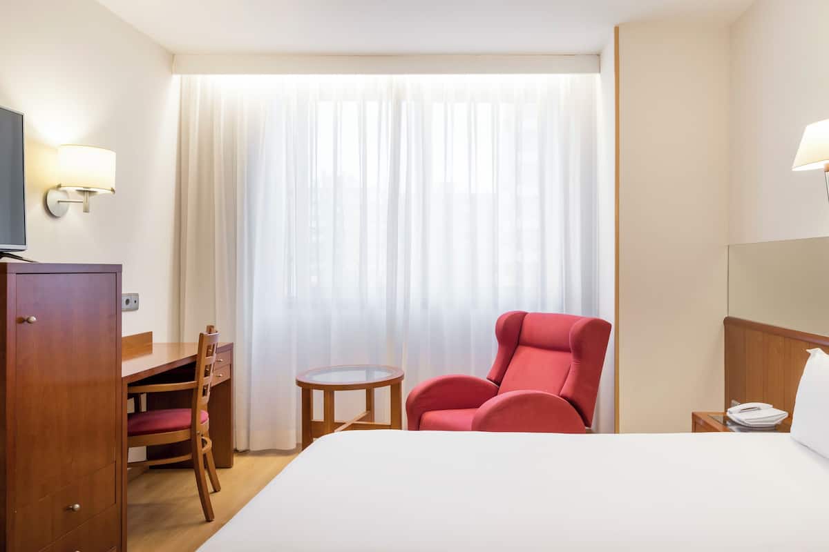 A hotel room with a bed, a desk with a chair, a red armchair, a small table, a television, and a mirror.