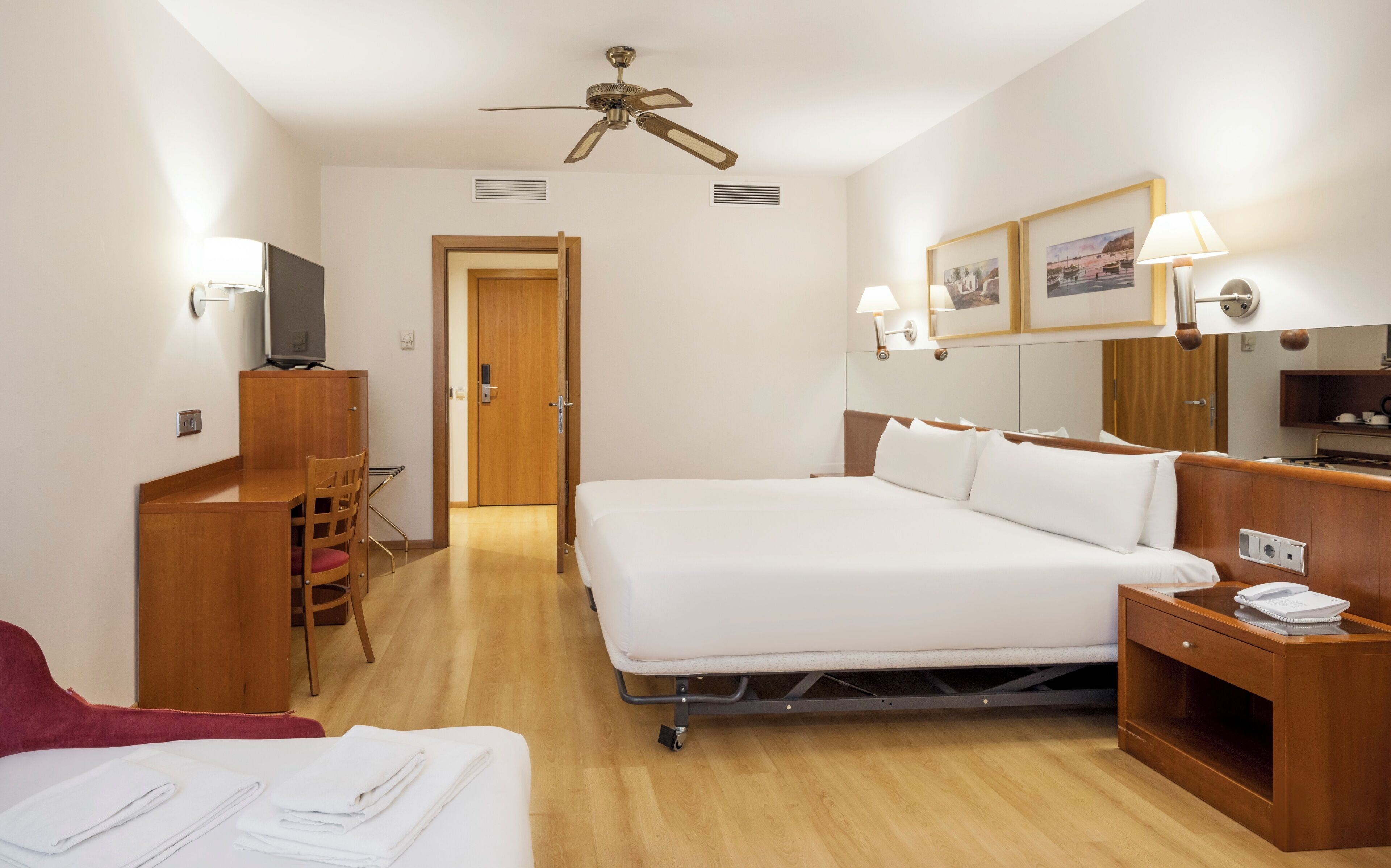 A hotel room with a bed, a desk, a chair, a TV, a ceiling fan, and a bathroom door.